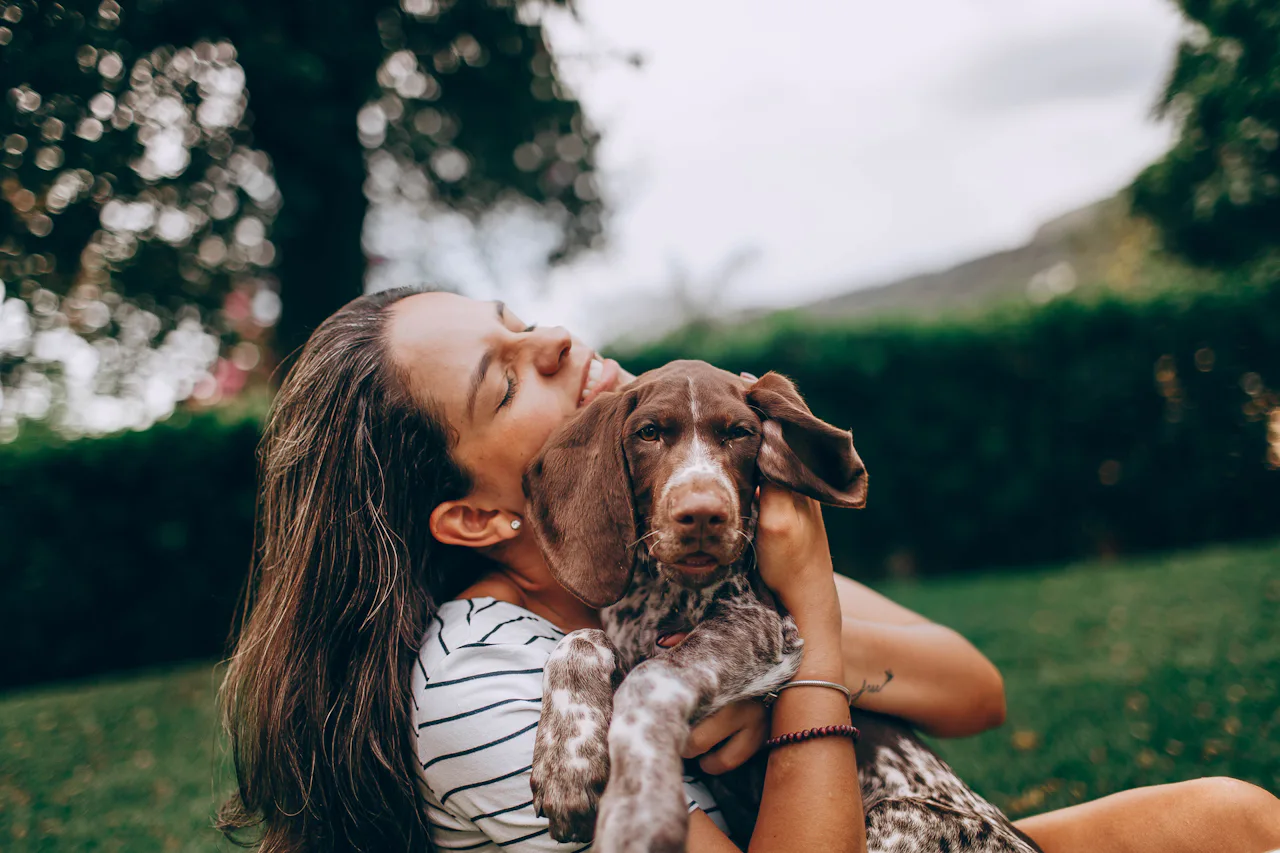 A woman sitting in the grass hugging her brown and white spotted pointer puppy